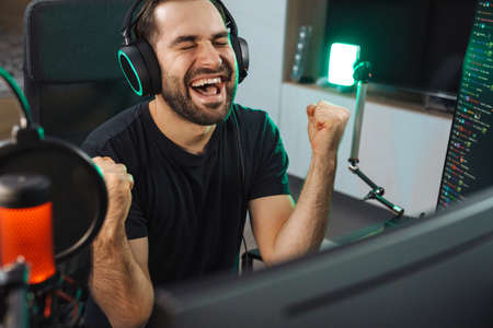 Happy young man gamer streamer in headphones playing on computer talking with players on chat in gaming competition celebrating winの写真素材