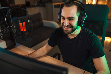 Happy young man gmaer streamer in headphones playing on computer talking with players on chat in...