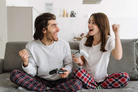 Young happy couple holding joysticks playing games on tv screen while looking at tv screen at home...