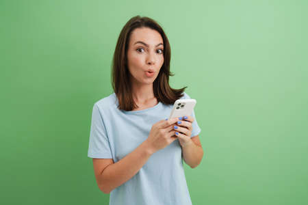 Young woman expressing surprise while using cellphone isolated over green background