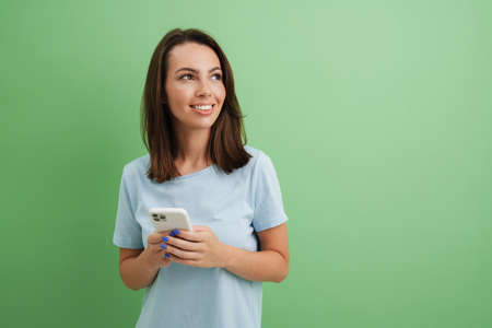 Young european woman in t-shirt smiling while using mobile phone isolated over green backgroundの写真素材