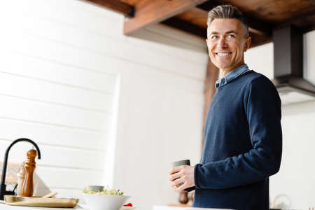 Happy mid aged white man holding cup standing in the kitchenの写真素材