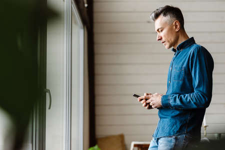 Mid aged casual man holding mobile phone standing indoors