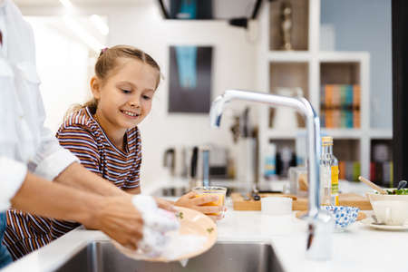White mother and daughter smiling while washing dishes together in kitchen at homeの写真素材
