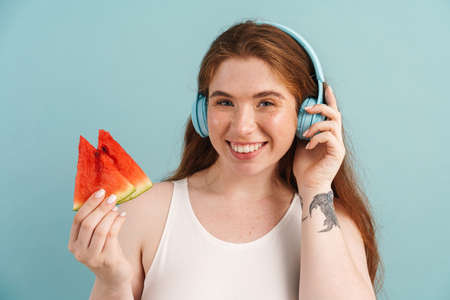 Young ginger woman in headphones posing with water melon isolated over blue backgroundの写真素材