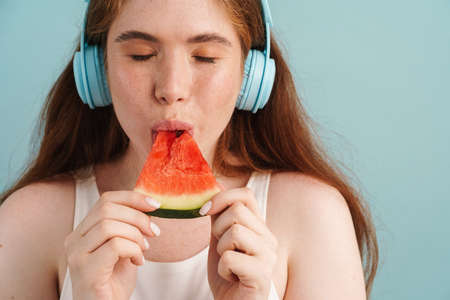 Young ginger woman in headphones eating water melon isolated over blue backgroundの写真素材