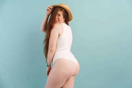 Young ginger woman in swimsuit smiling and looking at camera isolated over blue background