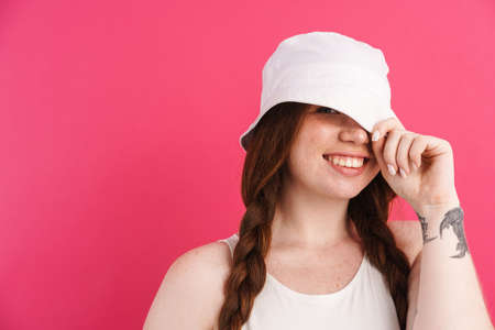 Young ginger woman with pigtails smiling and holding her panama isolated over pink backgroundの写真素材