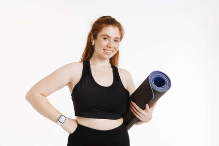 Smiling young plus size fitness woman in sportswear standing over white background carrying fitness matの写真素材