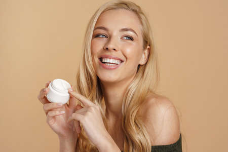 Smiling young blonde woman with long hair showing facial cream jar standing over beige backgroundの写真素材