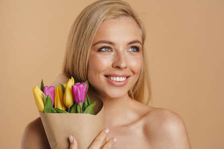 Happy young blonde white woman with long hair standing over beige background with bare shoulders holding bouquet of tulipsの写真素材