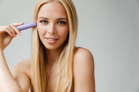Half-naked blonde woman looking at camera while showing mascara isolated over white background