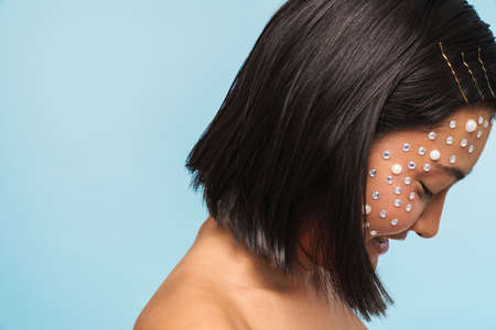 Beauty portrait of a young brunette asian woman with makeup facial gem decoration over blue wall backgroundの写真素材