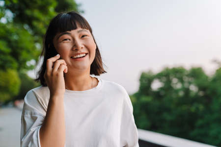 Smiling young asian woman walking in a green park talking on mobile phone close up