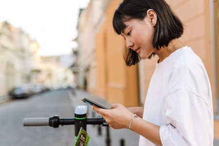 Smiling young asian woman using an app on smartphone to unlock a shared electric scooter on a city streetの写真素材