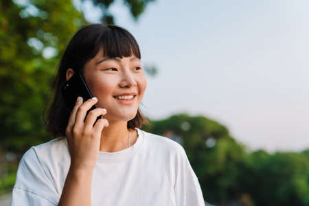 Smiling young asian woman walking in a green park talking on mobile phone close upの写真素材