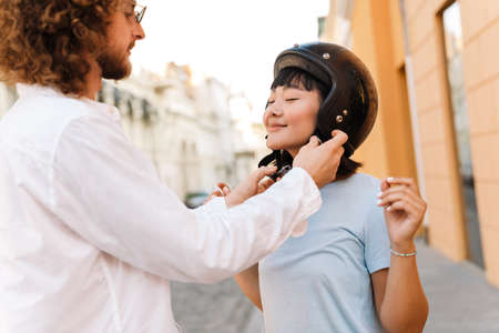 Smiling young man helping his asian girlfriend to put on her motorcycle helmet on a streetの写真素材