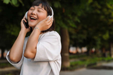 Smiling young asian woman walking in a green park enjoying listening to music with wireless headphones close upの写真素材