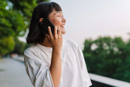 Smiling young asian woman walking in a green park talking on mobile phone close up