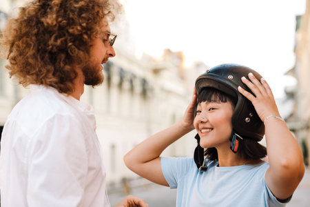 Smiling young man helping his asian girlfriend to put on her motorcycle helmet on a streetの写真素材