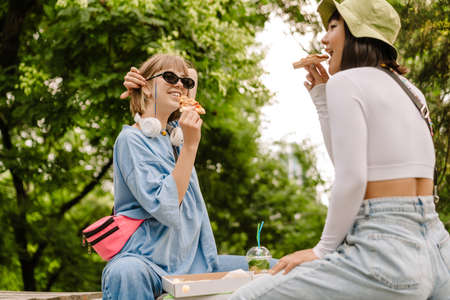 Multiracial two women smiling and eating pizza while resting in green parkの写真素材