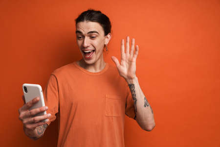 Portrait of a happy young white casual man with brown hair standing over red wall background holding mobile phone on a video call waving handの写真素材