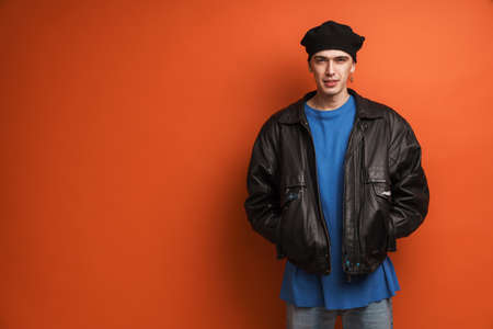 Young fashionable white man wearing leather jacket and beret standing posing over red backgroundの写真素材