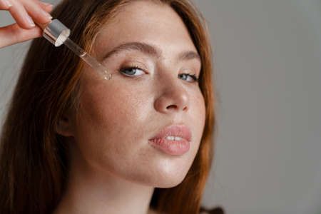 Portrait of a young white woman with long red hair standing over wall background, applying facial oilの写真素材