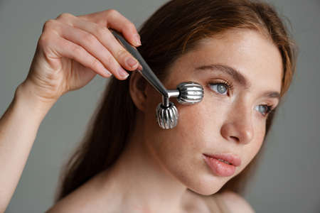 Young calm white woman massaging face with roller over gray background