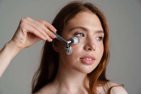 Young white woman using face massager while standing isolated on gray backgroundの写真素材
