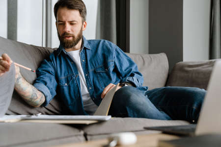 Bearded european man working with papers and laptop while sitting on sofa at homeの写真素材