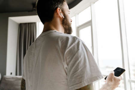 Back view of bearded european man in wireless earphones using mobile phone at homeの写真素材