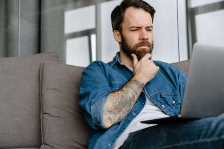 Bearded european man working with laptop while sitting on sofa at homeの写真素材