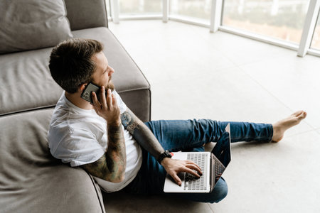 Bearded man talking on cellphone and working with laptop while sitting on floor at homeの写真素材