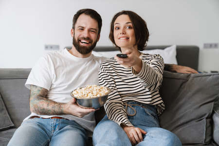 Happy young couple sitting on a couch at home watching tv holding remote control eating popcornの写真素材