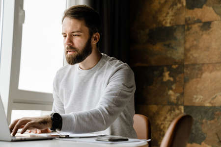 Bearded man working with laptop and papers while sitting at table indoors