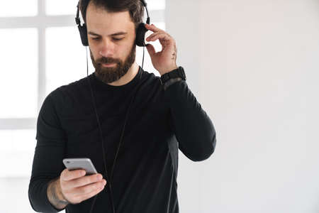 Young brunette bearded white man in sportswear standing indoors listening to music in headphones...
