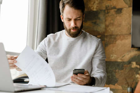 Bearded man using cellphone while working with laptop and papers indoors