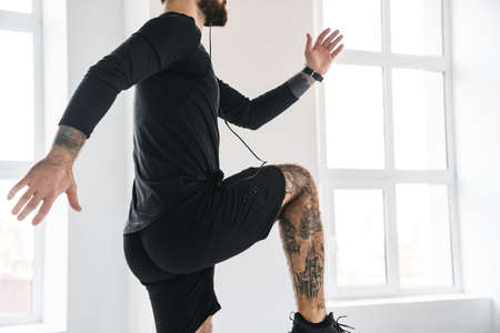 Young brunette bearded white man in sportswear exercising jumping indoors listening to music in...