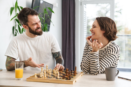 Happy young white couple playing chess sitting at the table at home talkingの写真素材