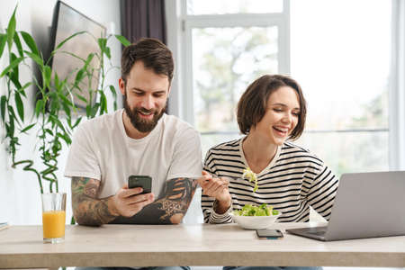 Smiling young white couple sitting at the table with laptop computer having breakfast holding mobile phoneの写真素材