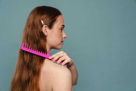 Half-naked young woman looking aside and combing her ginger hair isolated over blue background