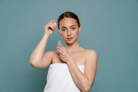 Portrait of a young healthy white woman holding bottle of facial oil serum at her face over blue wall backgroundの写真素材