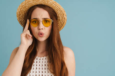 Young ginger woman in sunglasses expressing surprise at camera isolated over blue background
