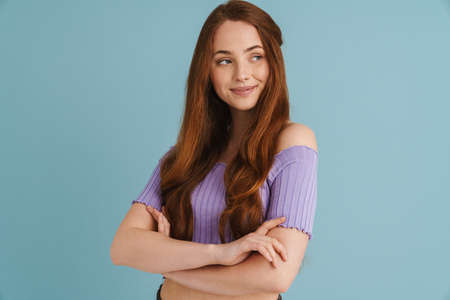Close up portrait of a smiling young casual woman with long red hair standing over blue wall...