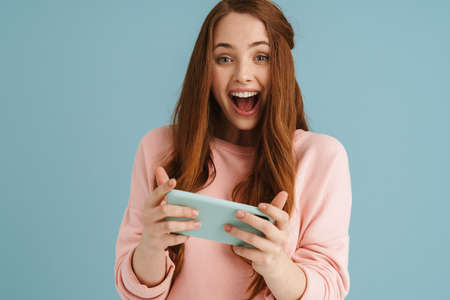 Young ginger woman laughing while using mobile phone isolated over blue background