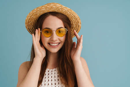 Young ginger woman in sunglasses smiling and looking at camera isolated over blue backgroundの写真素材