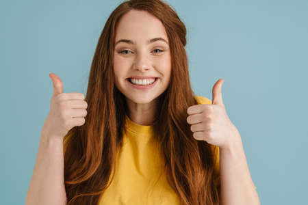 Young ginger woman smiling and showing thumbs up at camera isolated over blue background