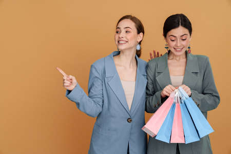 Young two women holding shopping bags and pointing finger aside isolated over yellow backgroundの写真素材