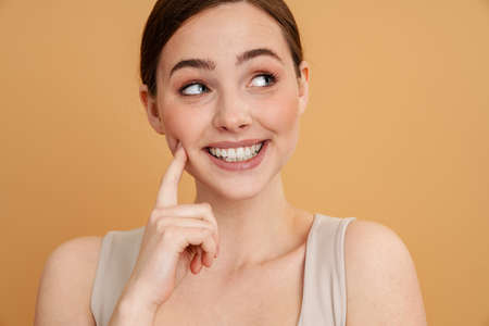 Close up portrait of a smiling young white red haired woman standing over beige background touching...
